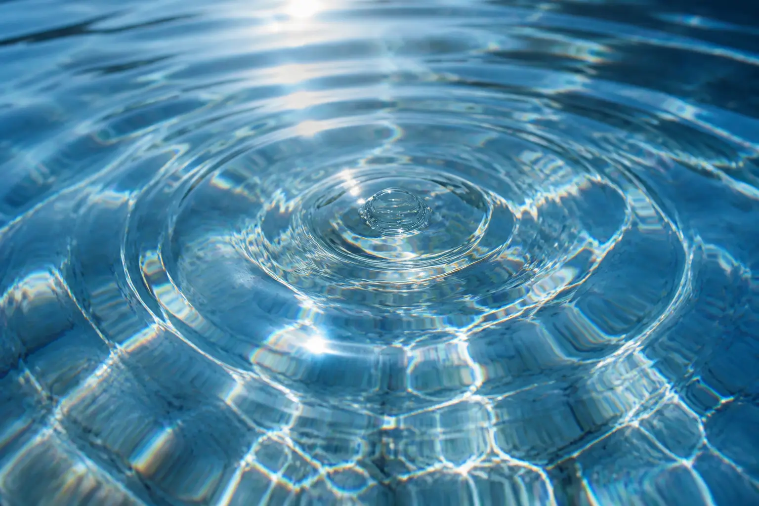 A close-up photograph looking down onto a clear, blue water surface, where a small, central water drop impact has created a series of precise, expanding concentric ripples. Bright sunlight illuminates the surface, creating shimmering highlights, sun glints, and complex refractive patterns across the circular ripple formation.