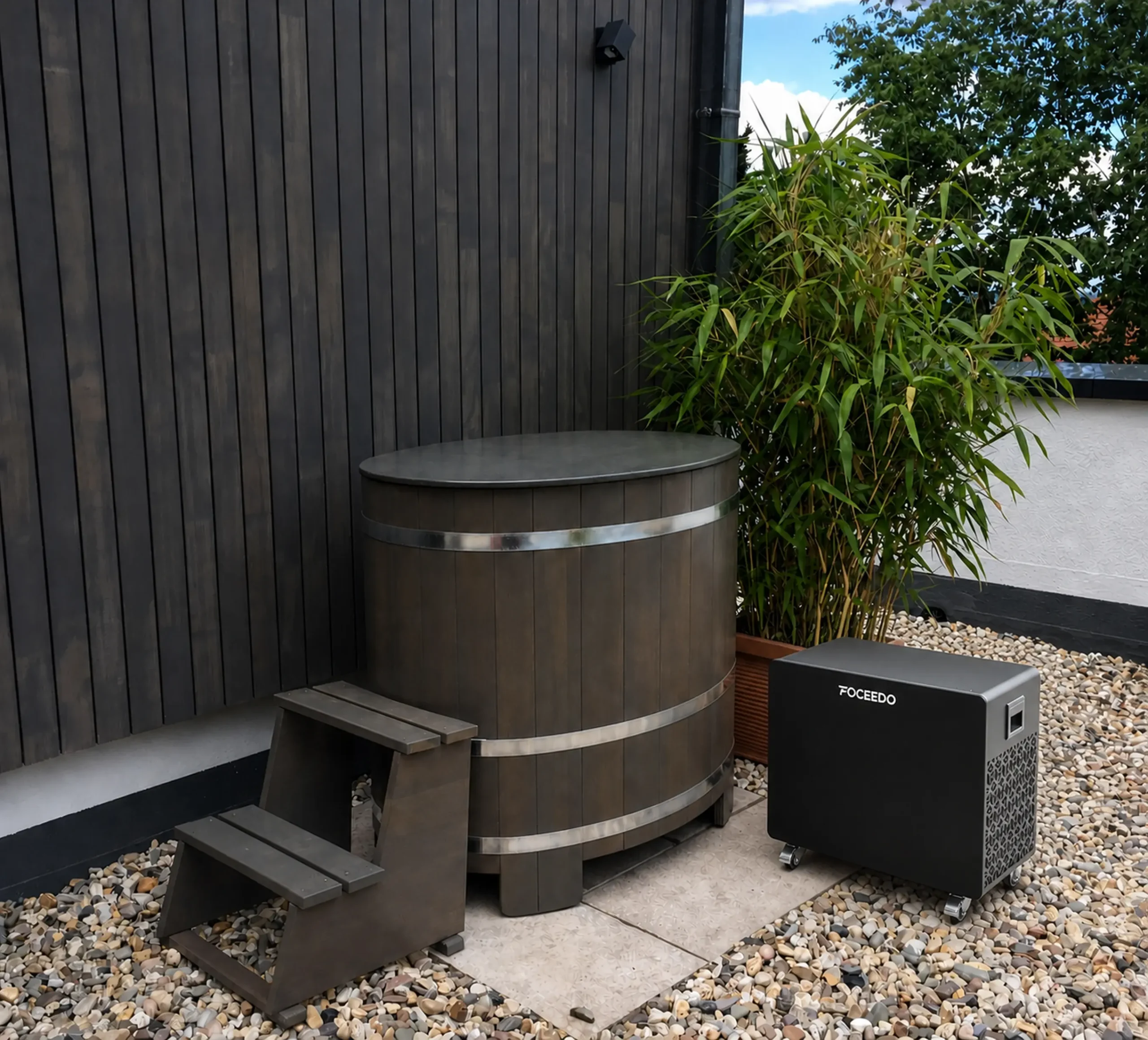 A Foceedo ice bath chiller placed next to a dark premium wooden tub on a pebble patio, showcasing a durable outdoor residential cold therapy setup.